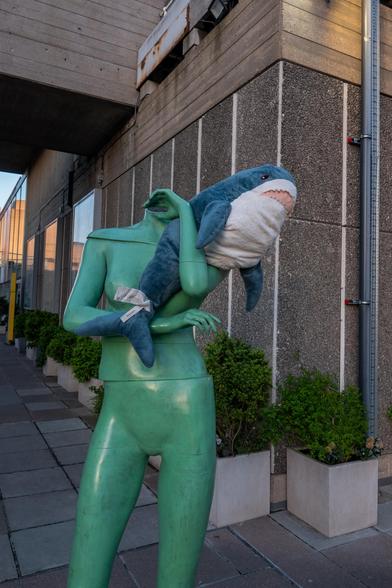 A blue and white Blåhaj Ikea shark is tucked between the arms of a green metal sculpture.