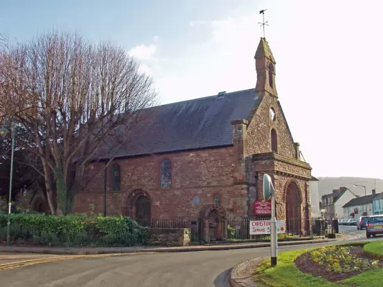 The image depicts a historic building, likely a church or community hall, set in a picturesque environment. The architecture features stone walls, arched windows, and a bell tower. There are trees nearby and the area appears to be well-maintained, with a roundabout and signs indicating community services. The overall setting suggests a quaint, small-town atmosphere.

Image Credits: Wikimedia / P Bly / CC BY-SA 3.0