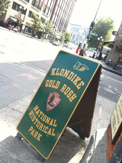 An a-frame sign on a city corner reads Klondike Gold Rush National Historic Park.