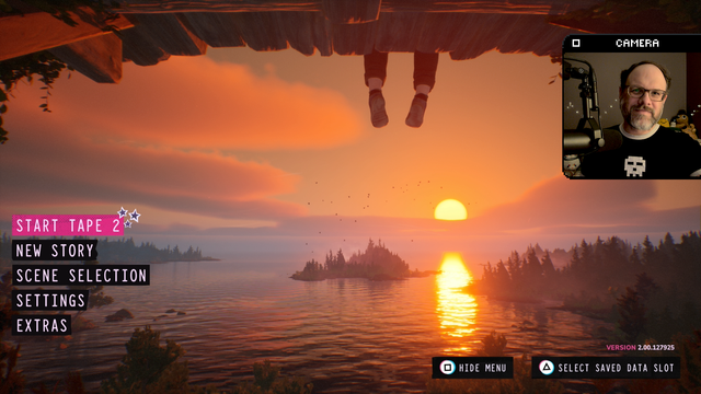 The title screen of Lost Records: Bloom and Rage. The camera is just under a wooden platform, with a single pair of legs dangling down. The sun is setting over a lake surrounded by trees. The game’s main menu is overlaid on the left side. The first option is highlighted in pink, with little stars drawn in the corner. It reads “start tape 2.”On the right is an inset image of me looking at the camera.