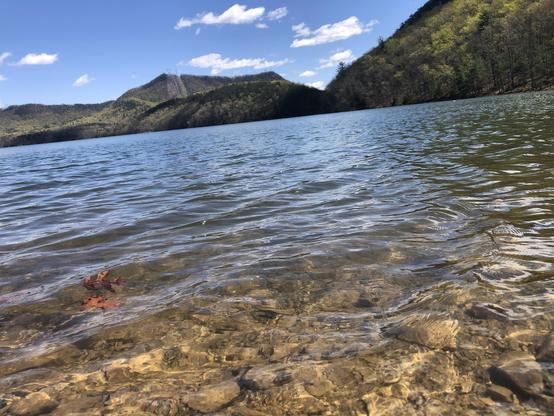 Small rocks under crystal clear ripples of lave water.  Across the lake are green mountains under a blue sky
