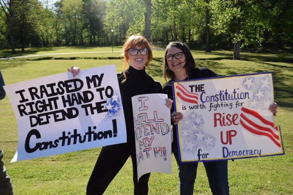 Two enthusiastic citizens holding signs. The first sign says "I raised my right hand to defend the Constitution ". The other sign says "The Constitution is worth fighting for. Rise up for democracy "