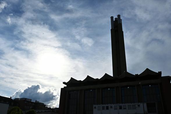This image shows a silhouette of a former power plant with distinctive features, including tall chimneys and a unique roof design. The scene is set against a backdrop of a cloudy sky, with the sun partially obscured, creating a dramatic contrast between light and shadow.