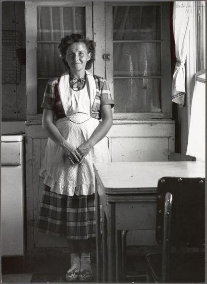 The image is a black and white photograph depicting an older woman standing in what appears to be a domestic kitchen or dining area. She wears traditional attire, including a plaid apron over a skirt with vertical stripes, which suggests the photo was taken during earlier times possibly mid-20th century. The setting features worn wooden cabinets, a simple desk chair, and a window draped partially by curtains. Her expression is one of contentment or amusement as she poses for the camera. No explicit actions are captured in this still image; however, her attire suggests domestic work, likely cooking or household duties within that era's social context.