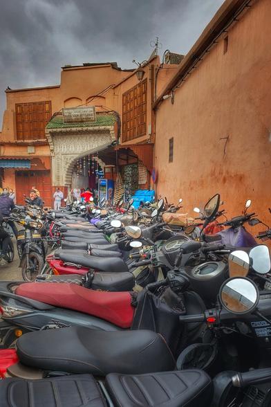 An eye-level shot captures a dense gathering of parked motorbikes and scooters in Marrakech, Morocco. The foreground is filled with numerous black and grey motorbikes, many with red or black seats, their handlebars and mirrors creating a complex network of lines. In the background, traditional Moroccan architecture rises, featuring peach-colored walls and arched doorways. One building has a green tiled roof and an ornate, light-colored facade with Arabic script. People can be seen walking in the background, adding a sense of everyday life to the scene under a cloudy sky.
