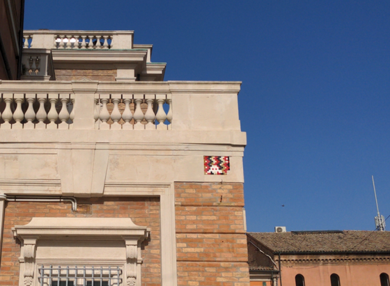 En haut d'un bâtiment néo-classique en brique et balustrade de pierre, sous un ciel bleu, un petit invader blanc, en mosaïque, sur fond de motif serpentant rouge, vert et blanc.