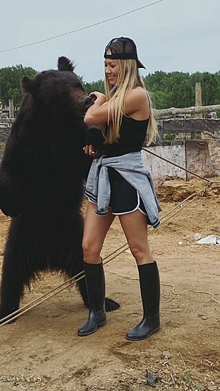 Veronica Dichka poses with her bear companion, Archie, a circus rescue.  She is dressed in rubber boots, running shorts, and a tank top, all black (the shorts piped in white) with a grey hoodie or sweater tied around her waist.  Her long blonde hair spills over her shoulders and across the arm she's holding across her chest while playing with the bear (who appears to be gnawing at it) as well as down her back.  A black baseball cap, bill to the rear, perches awkwardly on her head.

The bear is standing, one forepaw in her right hand, the other hanging down freely.
