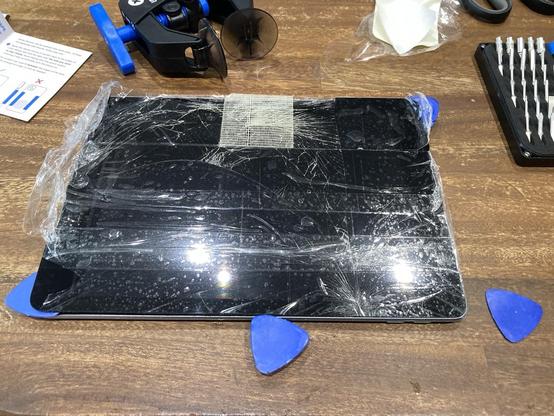 Ipad on wooden desk top. Packing tape is applied over entire front face. There are three blue spudger tools, resembling guitar picks, applied to front, left & right edges. One spudger waiting on the desk to be applied.