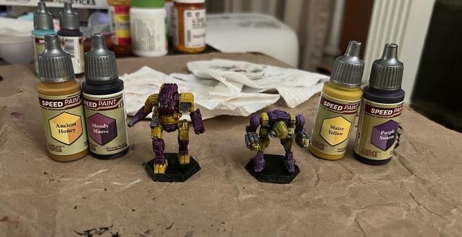 Two battle tech miniatures, both painted purple and yellow. 

All colors from Army Painter Speed Paints.

One on the left used Ancient Honey and Moody Mauve.

One on the right used Maize Yellow and Purple Swarm.