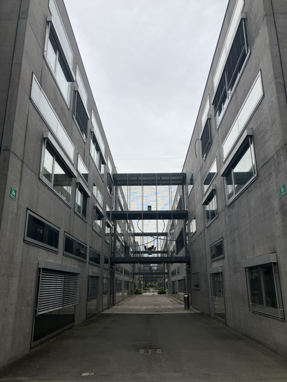 Picture of TU Graz campus, showing a walkway between buildings made of concrete and glass. Everything in the image is grey.