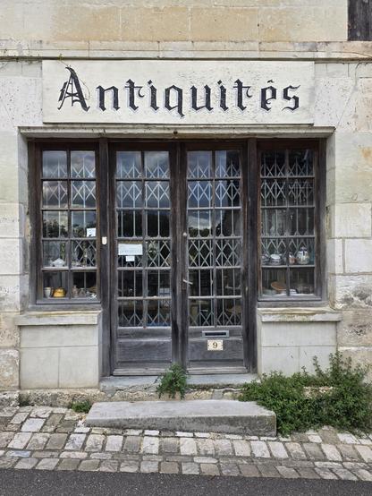 Phptograpb of a closed shop front. Dark wood double doors with a windor eitherside. Through the square panes of glass we can see metal shutters and crockerie. Above these is written the wird 'Antiquites' in black paint on sand stone in a medieval style