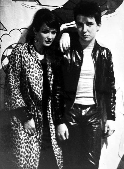 Grainy black and white photo of two teenagers. Jill is in a leopard print coat and rests her arm on Peter's shoulder. Peter is in shiny dark plastic jacket and trousers. Both look at the camera with expressions that say they know how cool they look.