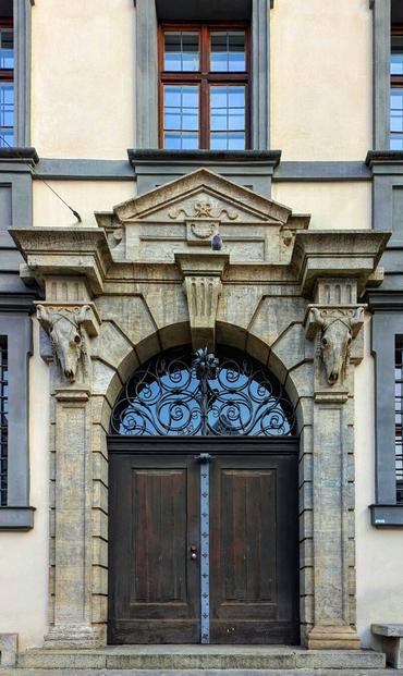 A close-up shows a grand, arched doorway of a historic stone building, the Stadtmetzg in Augsburg. Heavy, dark wooden double doors are framed by intricately carved grey stone. Above the arch, ornate ironwork features a central floral motif. Flanking the doorway are stone pilasters topped with weathered bull skull carvings. A decorative stone pediment sits above the architrave. The building's facade is a mix of pale yellow and grey stone.
