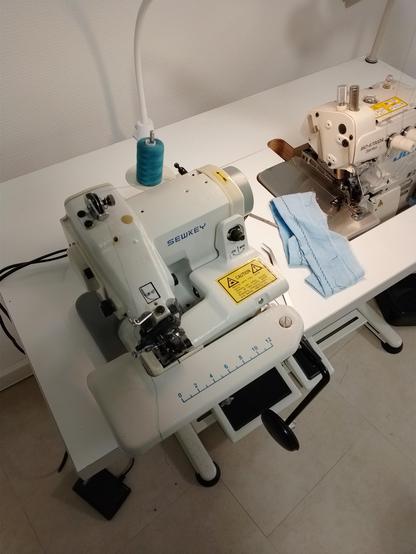 Eine kleine Blindstich-Nähmaschine auf einem Tisch. Es ist blaues Nähgarn eingefädelt. Daneben steht eine große Overblock-Nähmaschine.