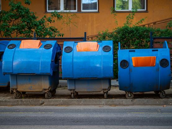 Vor einer orangefarbenen Hauswand eines Mietshauses drei Müllcontainer in kraftigem Blau, die Klappen unterschiedlich weit geöffnet. Auf den Klappen orangefarbene Akzente.