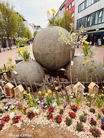 Nahaufnahme des österlich dekorierten Kugelbrunnens. Die großen Steinkugeln sind von bunten Frühlingsblumen, kleinen Insektenhotels und jungen Blütenbäumen umgeben. Ein harmonisches Zusammenspiel von Natur und urbanem Raum.