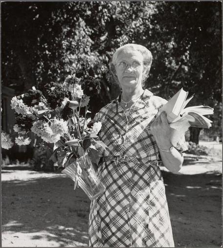 This black and white photograph features an elderly woman standing outdoors. She is holding a glass vase with flowers in her left hand, which appears to be filled with peonies or similar large blossoms. In her right hand, she holds several books loosely bound together by string. The woman wears glasses and has short hair that's pulled back from her face. Her attire includes a plaid dress with a pattern of squares predominantly featuring shades of black, white, brown, and red.
The background shows trees providing shade to the area where she stands, indicating this photo was likely taken in a garden or park setting during daytime. The lighting suggests it could be late morning or early afternoon based on the visible shadows cast by the woman's body and flowers within the vase. There is no direct sunlight shining directly onto her face, which adds to the softness of her facial features.
The overall composition conveys a sense of simplicity and contentment, possibly reflecting an interest in reading as well as gardening or nature appreciation. The image does not provide any explicit context for why she's posing with books; it might suggest that she is enjoying leisure time outdoors surrounded by flowers while engaging in the intellectual pursuit of literature.
This photograph was taken during a period when Dorothea Lange, known for her work documenting social issues and capturing moments from everyday life, captured images.  [...]