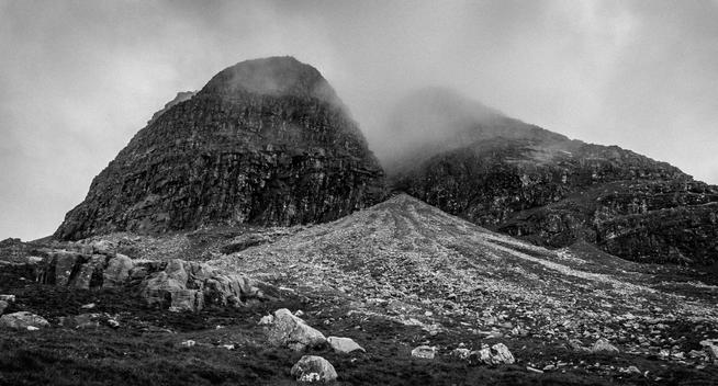 B&W image of clouds partially covering two prominent mountain peaks, with a sharp gap between them. In the gap, a flood of scree spills out covering most of the foreground of the photo. The sky is grey, in what is likely a rainy day.