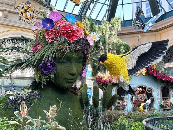 A sculpted bust of a woman, all green moss and leaves, water flowing from a cupped hand in front, a bird alighting on it for a drink.