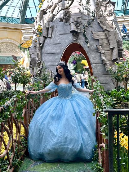 A woman in a lavish cindirella dress posing for a photo session in front of a large fairytale beehouse styled hut in a flower garden.