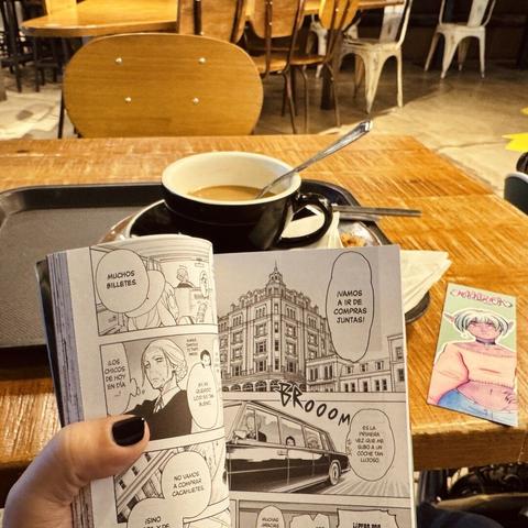 Bodegón de desayuno tras el manga abierto de Spy x Family. En la mesa está la bandeja con el café y, en un lado, el marcapáginas chiquito de una chica vampira de piel morada y pelo turquesa. O quizás es una orca :)