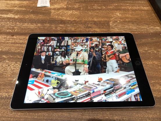 Ipad on wooden tabletop, now with front glass replaced. Bad Bunny’s NPR Tiny Desk concert is playing on the screen.