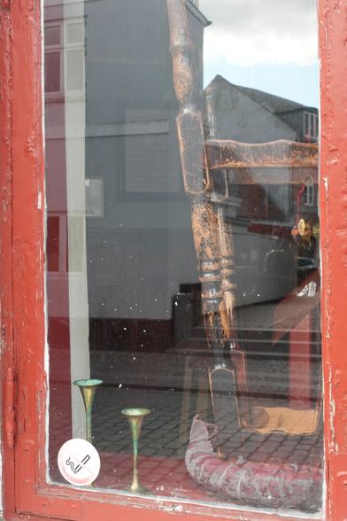Auf der rot lackcierten Fensterbank eines ebenfalls roten älteren Fensters steht innen ein alter Barhocker auf dem Kopf, von dessen rotem Lederbezug und dunkel lackierten Beinen bereits einige Farbe abgeblättert ist. danaben stehen zwei Messing-grüne Kerzenständer.  ein Tisch scheint im Hintergrund durch die Spiegelungen der gegenüberliegenden Gebäude hindurch.