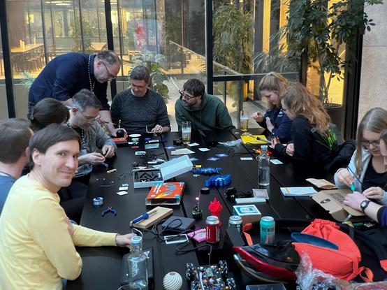 Eine Gruppe von Menschen sitzt an einem großen Tisch mit verschiedenem Lötmaterial (Hardware und allgemeines Bastelequipment).
