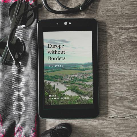 europe without borders a history by isaac stanley-becker book, portraying a very classically european view of a river with a bridge across it, leading to a town, and a beautiful, valley-and-hills forested horizon. black framed LG tablet is placed on a dark grey wooden table, with various dark items strewn around, such as arasaka facemask, round black shades, earphones. review by reading nosferatu, image by vakaris aka wannabekingpin.