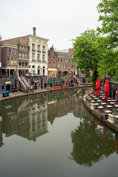 The image depicts a serene canal scene in a European city, likely in the Netherlands, characterized by its historic architecture and waterways. The foreground features a calm canal with reflections of the surrounding buildings and trees. On the right side, there is a row of outdoor café tables with red umbrellas, suggesting a leisurely atmosphere. The buildings along the canal are multi-storied, with a mix of brick and stone facades, and some have ornate details and large windows. A tree with green leaves adds a touch of nature to the urban setting. The sky is overcast, contributing to the tranquil ambiance. The scene is devoid of people, emphasizing the quietness of the moment.

Provided by @altbot, generated privately and locally using Ovis2-8B

🌱 Energy used: 0.177 Wh
