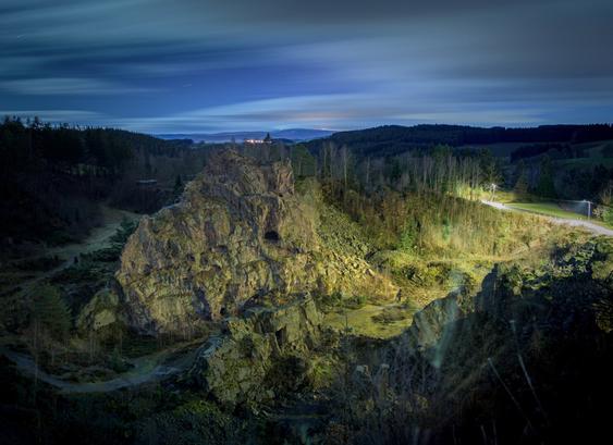 DE: Nächtlicher Blick auf einen großen zerklüfteten Granitbrocken, der als Art Mahnmal aus einem Kessel herausguckt. Von Westen bescheinen Straßenlaternen die Felswand, im Osten beleuchtet der bedeckte Vollmond die Umgebung. Kilometerweit im Hintergrund sind weitere Berge und Orte des Erzgebirges zu entdecken. Durch die fast 4,5 Minuten Belichtung sind die Stern zu Spuren und Wolken zu einer diffusen Fläche verwandelt.

EN: Night-time view of a large, jagged chunk of granite peeking out of a cauldron as a kind of memorial. Streetlights illuminate the rock face from the west, while the overcast full moon lights up the surroundings in the east. Further mountains and towns in the Ore Mountains can be seen for kilometres in the background. The almost 4.5-minute exposure has turned the stars into trails and the clouds into a diffuse surface.