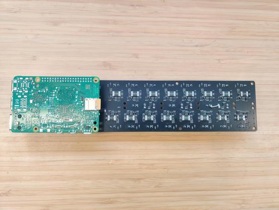 A long thin black PCB with 16 white connectors, rear side, with the underside of a Raspberry Pi visible on the left