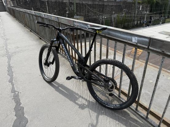 Ein schwarzes MTB lehnt an einem Geländer einer Brücke. Hinten unterm Sattel sieht man gelbe Kabel um das Dattelgestell gewickelt.