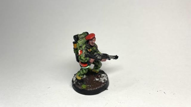 a miniature in green forest camo, a large square backpack with a lasgun attached via thick cables. They’re wearing a red beret.