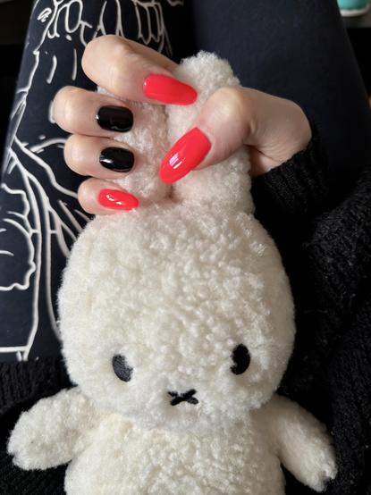 I’m holding my Miffy plushie by the ears with my right hand, showing my nails. My middle and ring fingers are short and black, the others are very long and a very bright red-orangey color