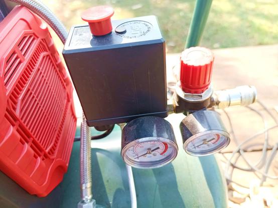 Close-up of a black plastic box with red knob on top, which houses a pressure switch for switching the compressor on when the air pressure drops below a threshold, and off again when it reaches another one that should be around 8 bar. There's also a red regulator knob and two gauges. On the right is a standard air hose coupling, on the left a red plastic cover for the pump.