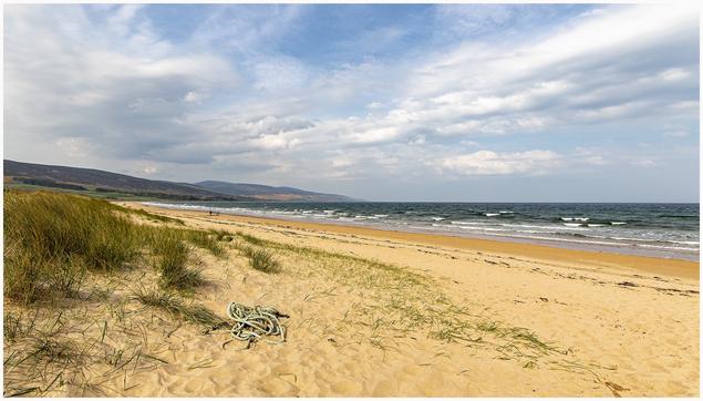 A sandy beach with rolling waves and a cloudy sky. In the foreground, there is a bundle of rope alongside patches of grass. A few people can be seen walking near the water's edge in the distance, with hills visible in the background.
