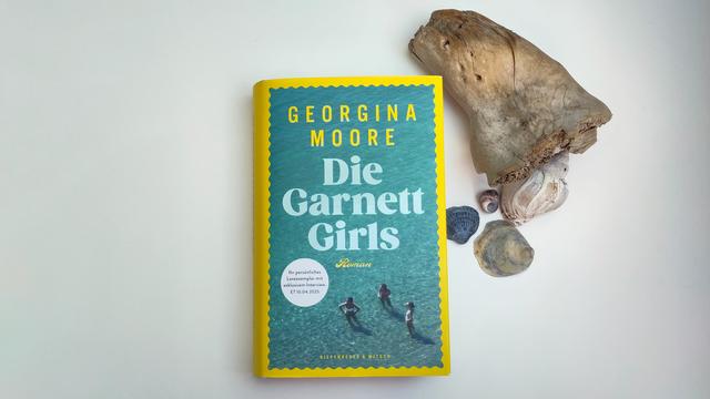 Das Buch liegt auf einer hellen Fläche. Oben rechts ist ein Stück Treibholz und darunter vier Muscheln zu sehen. Das Bild ist mit roten Linien umrahmt. Das Buchcover ist in der Grundfarbe leuchtend gelb. Das Cover zeigt eine Fotografie, die mit einem Wellenschnitt umrandet ist. Das Foto ist eine Aufnahme von oben, es zeigt drei junge Frauen in Badebekleidung, die im seichten türkisfarbenen Wasser stehen. Oben steht in gelben Versalien der Autorinnenname und darunter dreizeilig in hellblauer Serifenschrift der Titel „Die Garnett Girls“. In einer kleinen gelben Schmuckschrift folgt der Zusatz „Roman“. Links ist ein grauer runder Aufdruck mit dem schwarzen Text: „Ihr persönliches Leseexemplar mit exklusivem Interview. ET 10.04.2025“. Mittig am unteren Rand steht in kleinen gelben Versalien der Verlagsname.