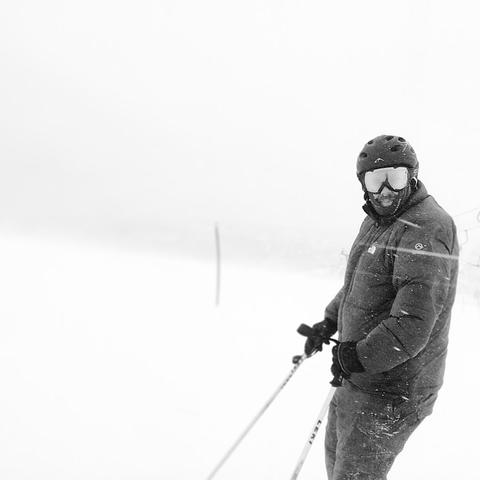 Skier in winter gear standing on a snowy slope during a blizzard. He wears a helmet, ski goggles, and a padded jacket. Snow is visibly blowing across the frame. A thin pole is faintly visible in the background, marking part of the ski route. Visibility is low due to heavy snowfall.