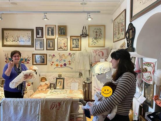 A photo of a room with white walls and lots of things on display: the walls are covered in framed art and photos, there's a table with embroideries and knick-knacks, and it gives off a vibe of a lovingly cluttered home. The lighting above gives it away as a museum gallery. We see an older woman with short dark hair, a bluey-purple top, and glasses holding a swaddled baby doll in the background. She's talking to the woman in the foreground, who has long dark hair and a striped top, and is holding a baby with a smiley face over her actual face.