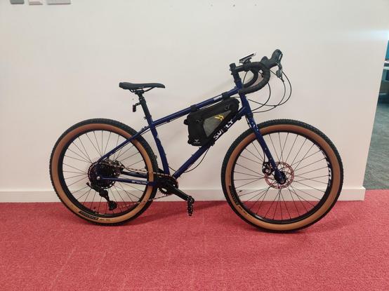 A shiny new bike leaving against a white wall on a red carpet in an office. 
It's a blue hardtail frame with drop handlebars and rigid forks. The frame bag in the middle looks tiny, and I'll be forced to buy a new one. 
The tires are 2.5 inch mountain bike tires with tan sidewalls.