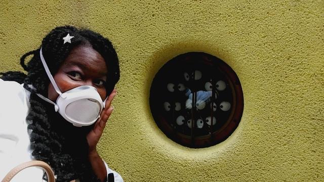 Jay is wearing a white Flo mask as she holds her hand to her cheek, looking alarmed while next to a small round window with bars on it, where many wide eyed soot sprites are being held captive inside a building with yellow walls at the Ghibli Museum in Tokyo, Japan.