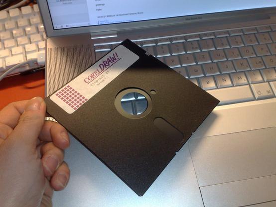 a left hand holder a 5,25" floppy disk. The label on the disk reads CorelDRAW! In the background an old Apple PowerBook with its silver keycaps.