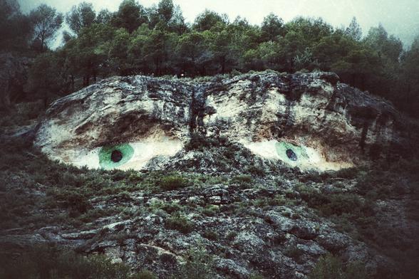 A wide shot of a rocky hillside with two large, green eyes with black pupils that are painted onto the natural rock formation, surrounded by dark green vegetation and trees in the background under an overcast sky.