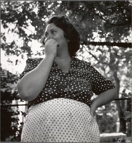 The image is a black and white photograph depicting an older woman standing outdoors. She appears to be in her late forties or early fifties, with medium-length dark hair styled slightly away from the face. The subject's complexion suggests she might have been of Jewish descent.

She wears a sleeveless blouse adorned with large polka dots and a full-skirted skirt featuring small white circles on a pale background. Her right hand is gently resting on her cheek, while her left arm hangs by her side. She seems to be in deep thought or contemplation as she gazes off into the distance.

The backdrop features an outdoor setting with trees providing shade, and it appears to be daylight given the natural lighting of the scene. The photo has a candid quality, capturing a moment that feels intimate yet universal.

In terms of context provided by the alt text, this image is connected to Dorothea Lange's work during her travels in America during 1858 when she documented various communities and their lifestyles. Bessie seems to be part of such documentation as one of ten families settled on an isolated ox-cart trail.

The photograph has a signature or inscription at the bottom right corner, indicating it may have been taken by Dorothea Lange herself or someone associated with her work during that era.