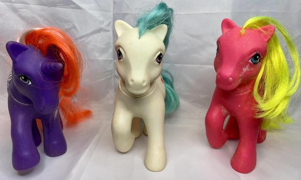 Three colorful toy ponies are positioned side by side against a white background. The left pony is purple with an orange mane and a unicorn horn. The middle pony is white with a teal mane. The right pony is pink with a yellow mane.