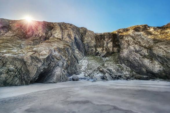 A wide shot of a sandy beach leading to large, rocky cliffs under a clear blue sky with the sun shining over the top of the cliffs. At the bottom of the cliffs is a small, dark cave entrance.