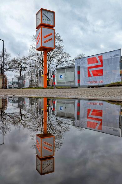 Das Bild zeigt eine Baustelle mit einem markanten, orangefarbenen Uhrenturm im Vordergrund. Der Turm hat zwei quadratische Uhren, die die Uhrzeit auf analogen Zifferblättern anzeigen. Unterhalb der Uhren befindet sich ein quadratisches Schild mit der Aufschrift „Zu vermieten 089/4809980“ in roter Schrift. Auf dem Boden ist eine große Pfütze, in der sich der Uhrenturm sowie umliegende Bäume und Bauzäune spiegeln. Im Hintergrund erkennt man einen Bauzaun, auf dem ein großes Banner mit der Aufschrift „Mayr“ und „mmibau.de“ in roter Schrift befestigt ist. Das Wetter wirkt bewölkt, und es scheint, dass es vor Kurzem geregnet hat.