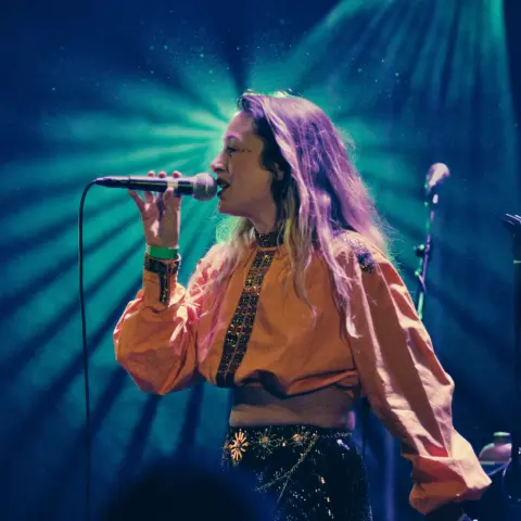 Photo de la chanteuse Luiza, en train de chanter dans un micro, avec une lumières a rayons en fond et de la poussière visible au dessus.