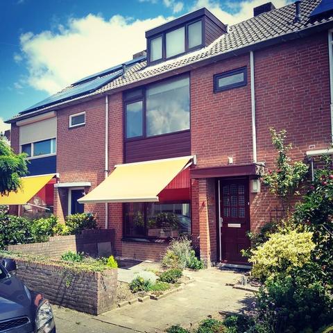 Voorgevel van een huis uit een 70-er jaren nieuwbouwstraat. De voortuintjes er omheen zijn vol met groene planten en bloemen, maar dit huis heeft tegels met wat grind en een paar kleine plantjes. Het is huisnummer 4. Een oranje zonnescherm is naar beneden. Het houtwerk van deur en kozijnen en dakkapel is bruin.